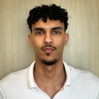 Jeune homme avec cheveux bouclés noirs et chemise blanche devant un mur clair.