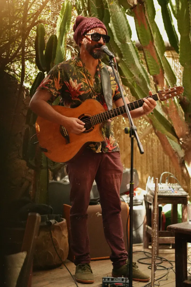 A man wearing sunglasses, a patterned shirt, and a beanie plays an acoustic guitar and sings into a microphone surrounded by large cactus plants in a warm, indoor setting.