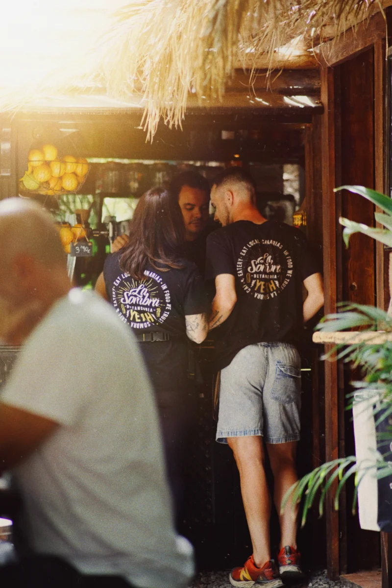 Two people wearing black t-shirts with 'La Sombra Betancuria' logo stand inside a cozy, rustic bar decorated with hanging oranges and wooden elements, while a person in a white shirt is blurred in the foreground.