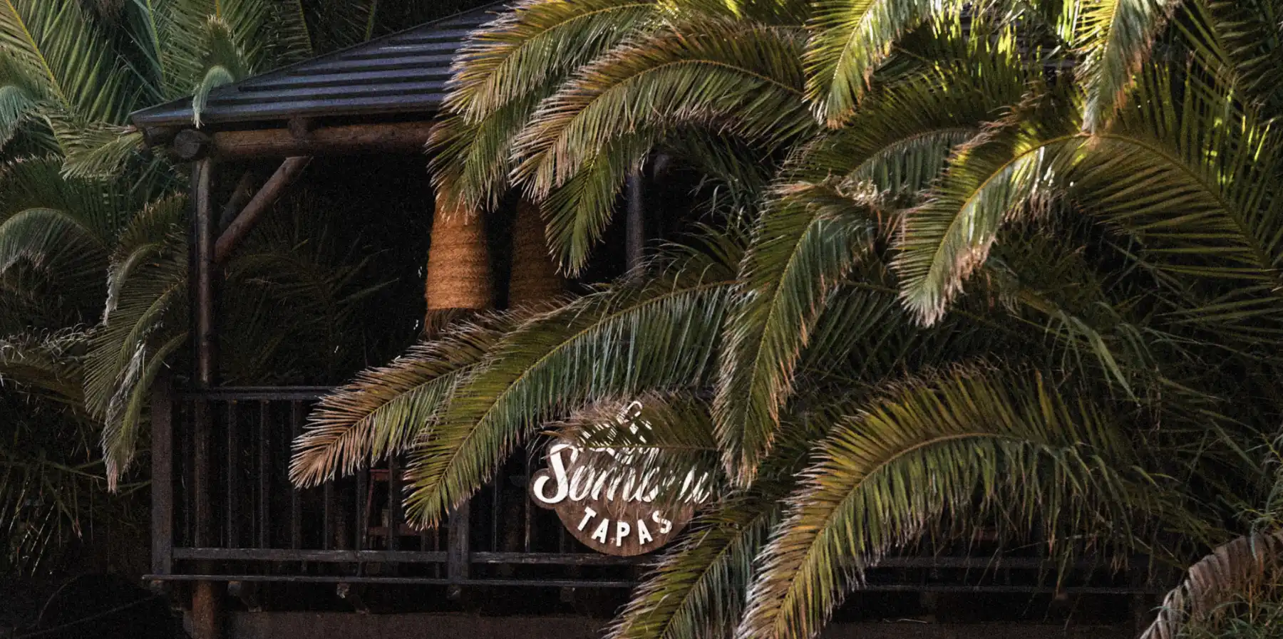A cozy outdoor wooden balcony partially hidden by large palm tree leaves, with a hanging sign that reads 'La Sombra'.