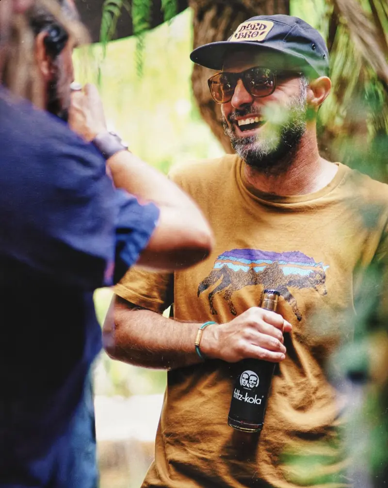 A man wearing sunglasses, a black cap, and a brown t-shirt with a wolf graphic is holding a bottle of Fritz-Kola and smiling at another person.