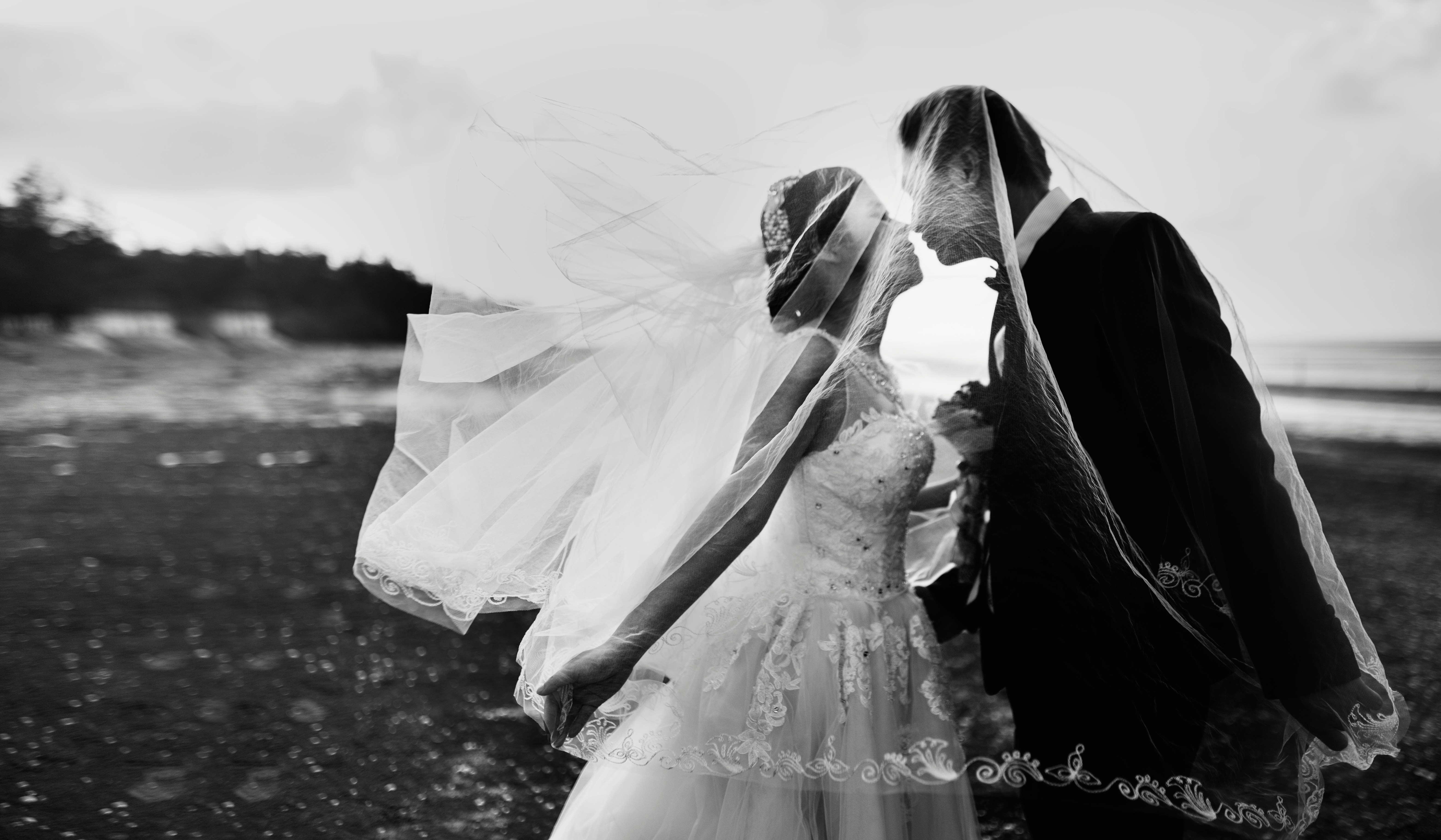 Bride and groom sharing a kiss under the bride's flowing veil on a beach at sunset.