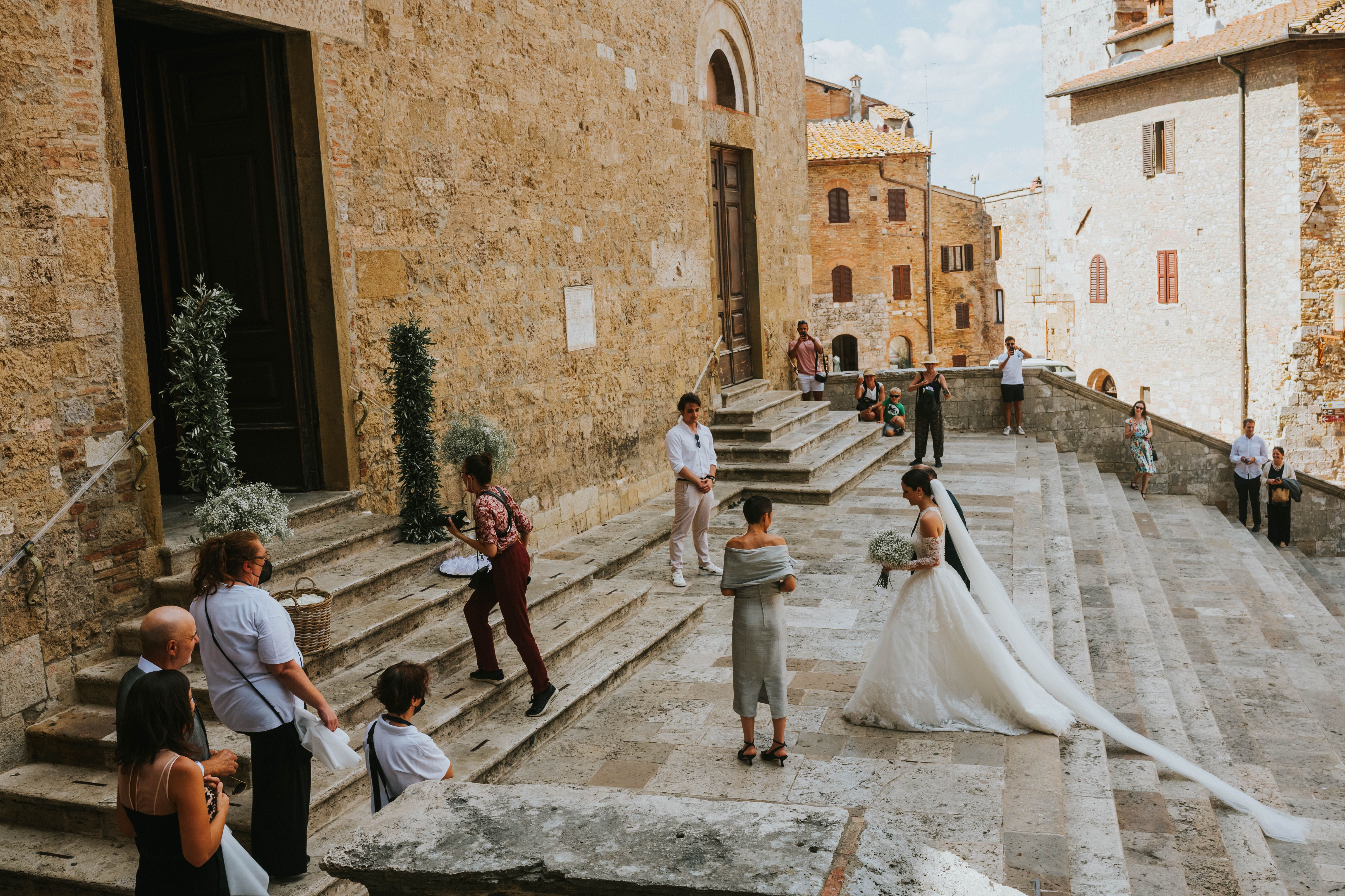 Bride in long veil holds bouquet on stone steps with guests and photographers nearby.