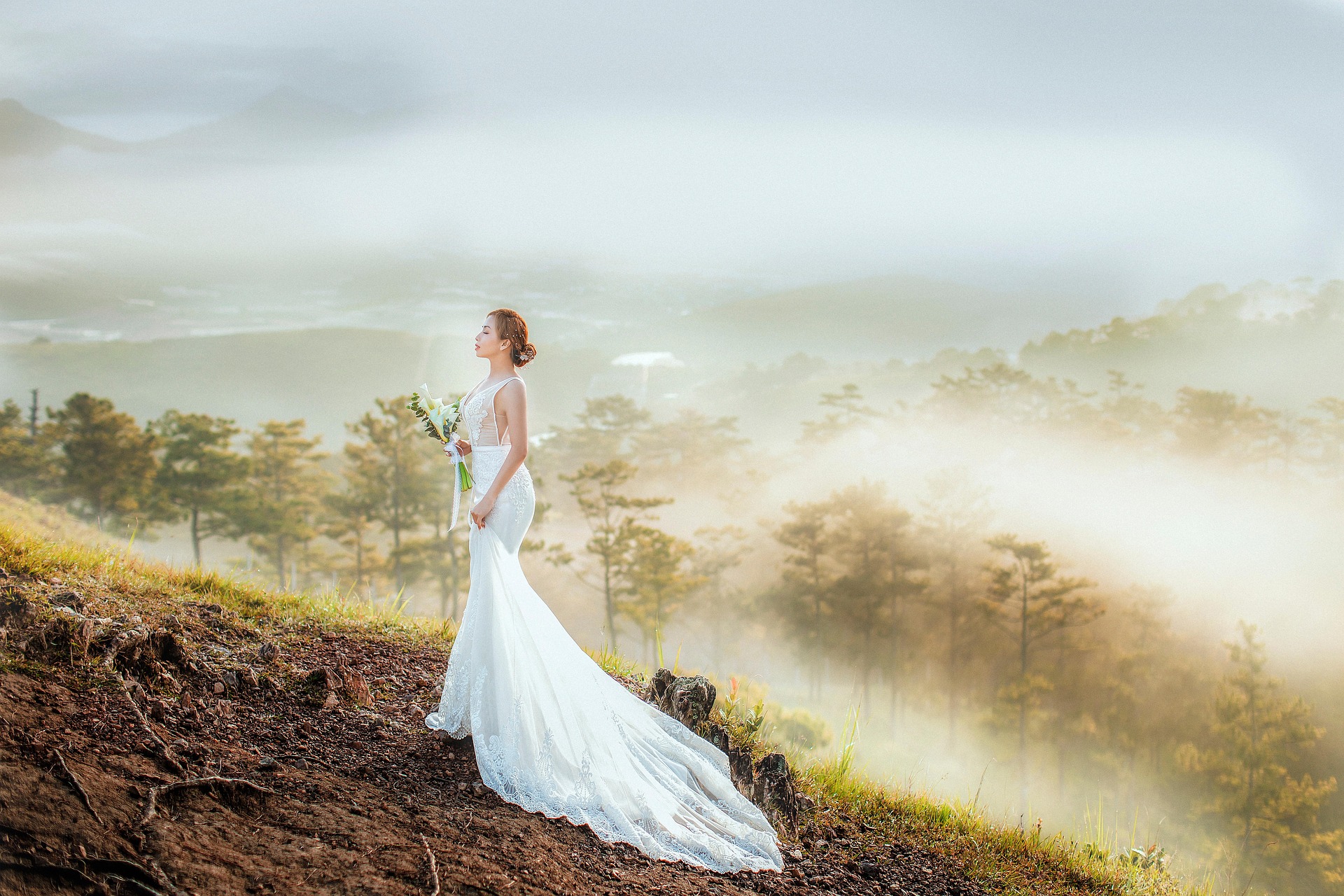Bride in white wedding dress holding bouquet, standing on grassy hill with foggy forest in background.