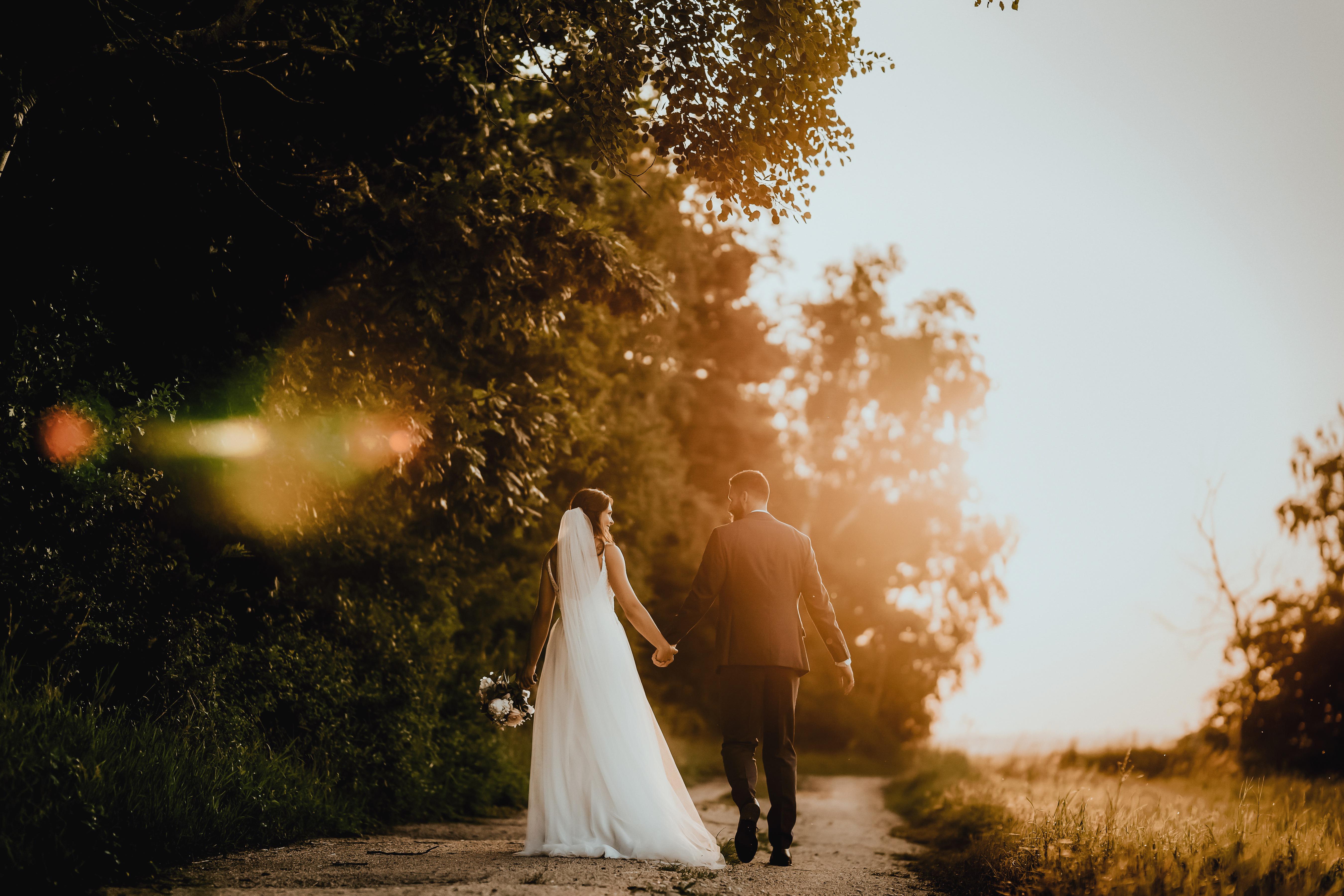 Bride in white dress and groom in suit walking hand in hand on a tree-lined path at sunset.