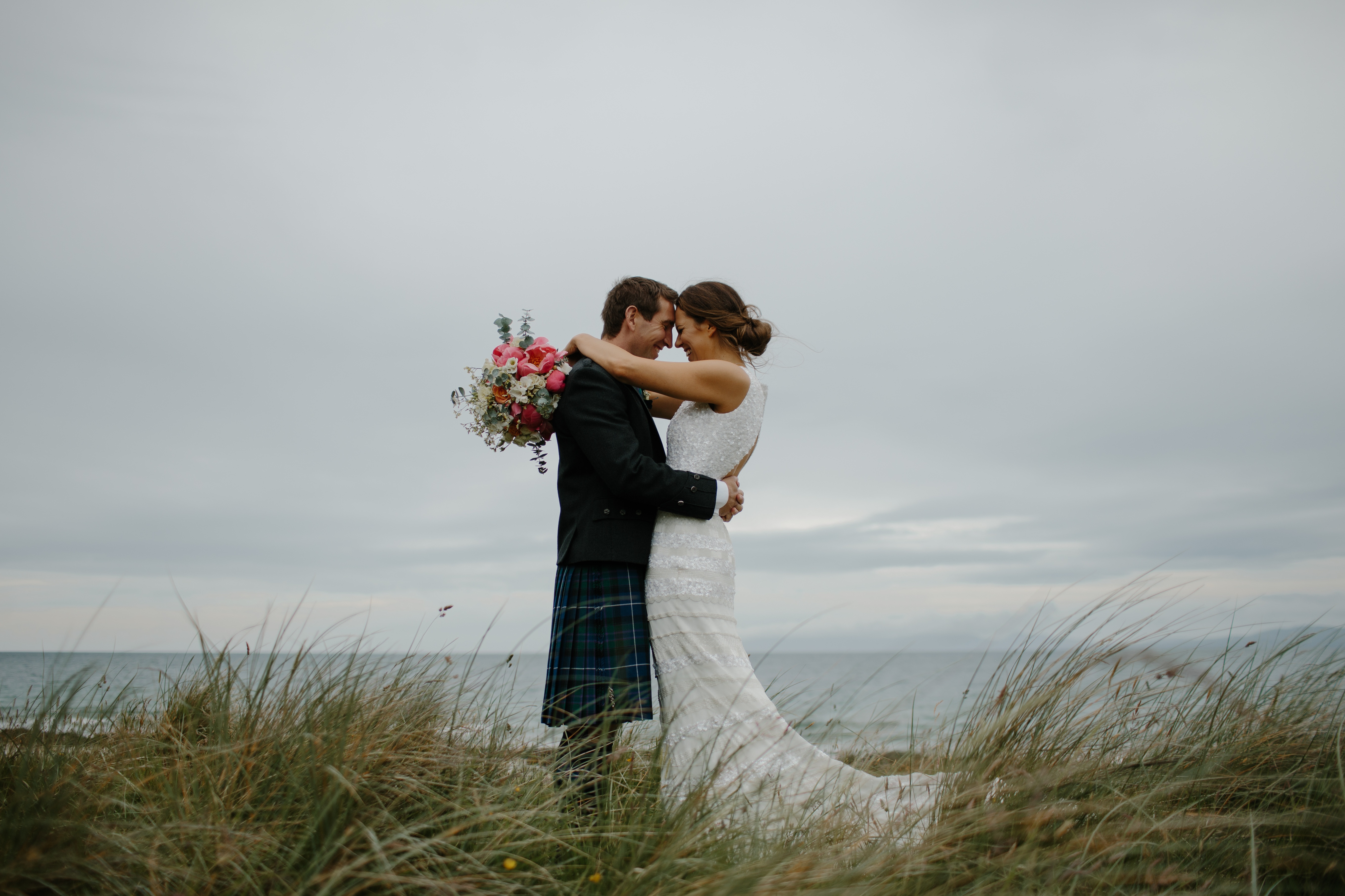 Bride and groom in a Scottish kilt embracing on a grassy shore with ocean in the background.