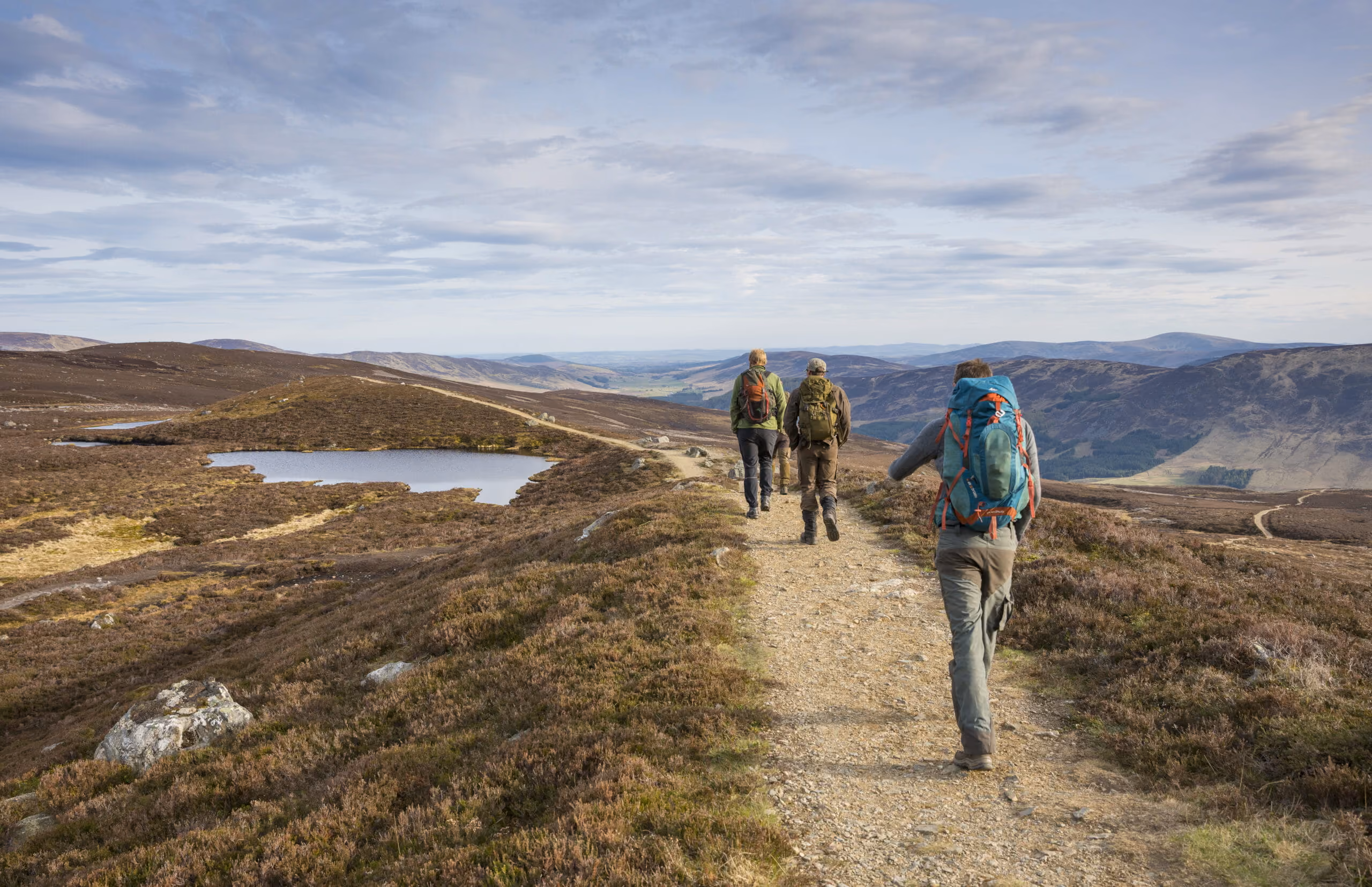 Three hikers with backpacks walking on a dirt trail in a mountainous landscape with lakes.
