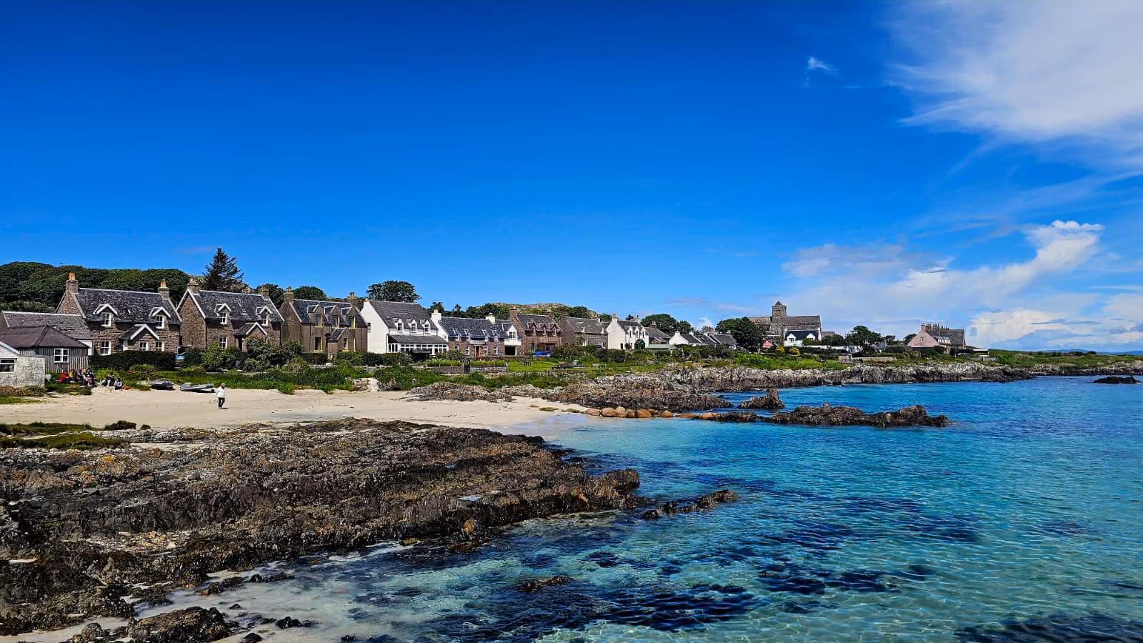Coastal village with stone houses, rocky shoreline, sandy beach, and clear blue sea under a sunny sky.