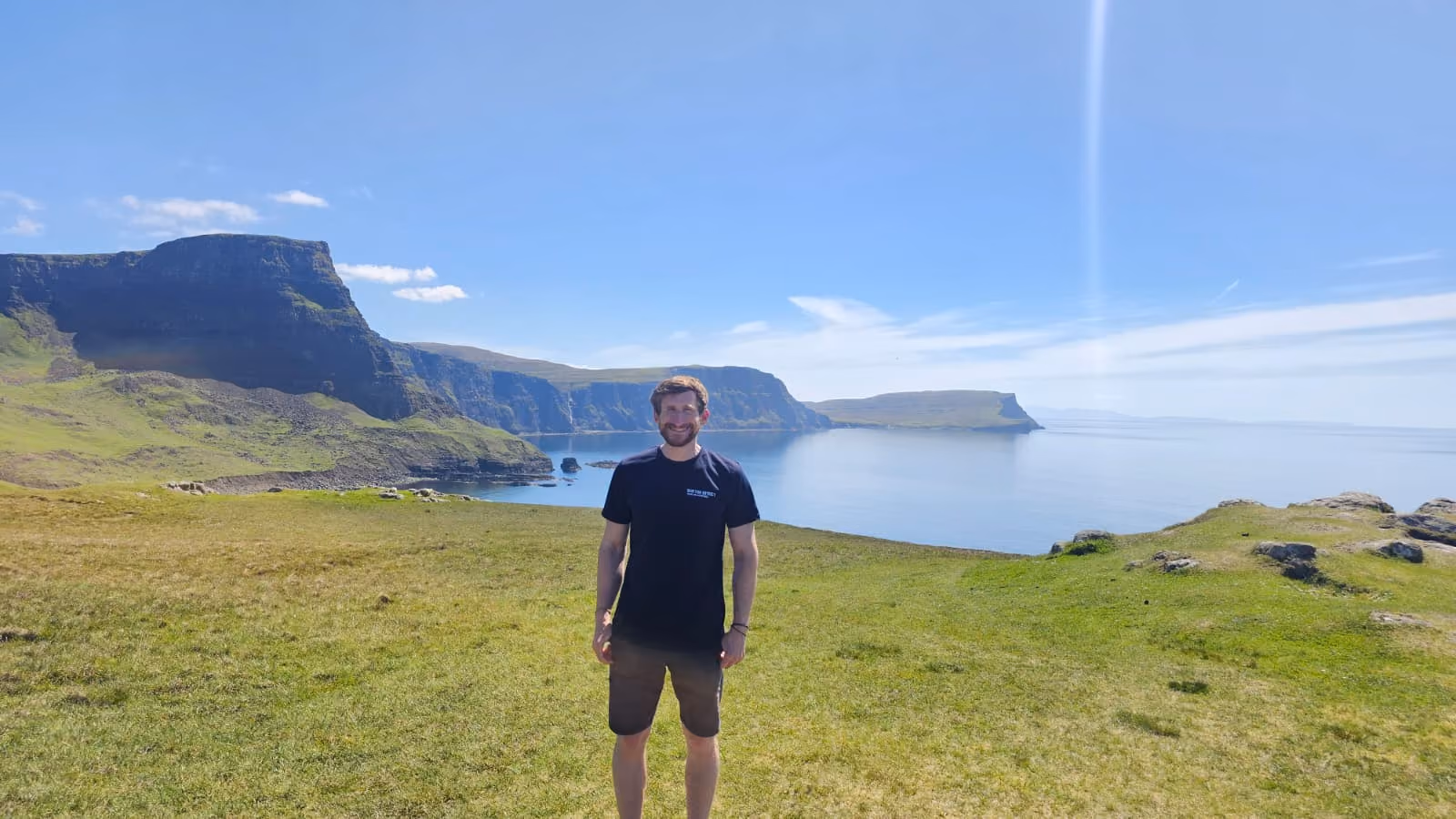 Smiling man in black shirt and shorts standing on green grass with cliffs and calm sea behind him.