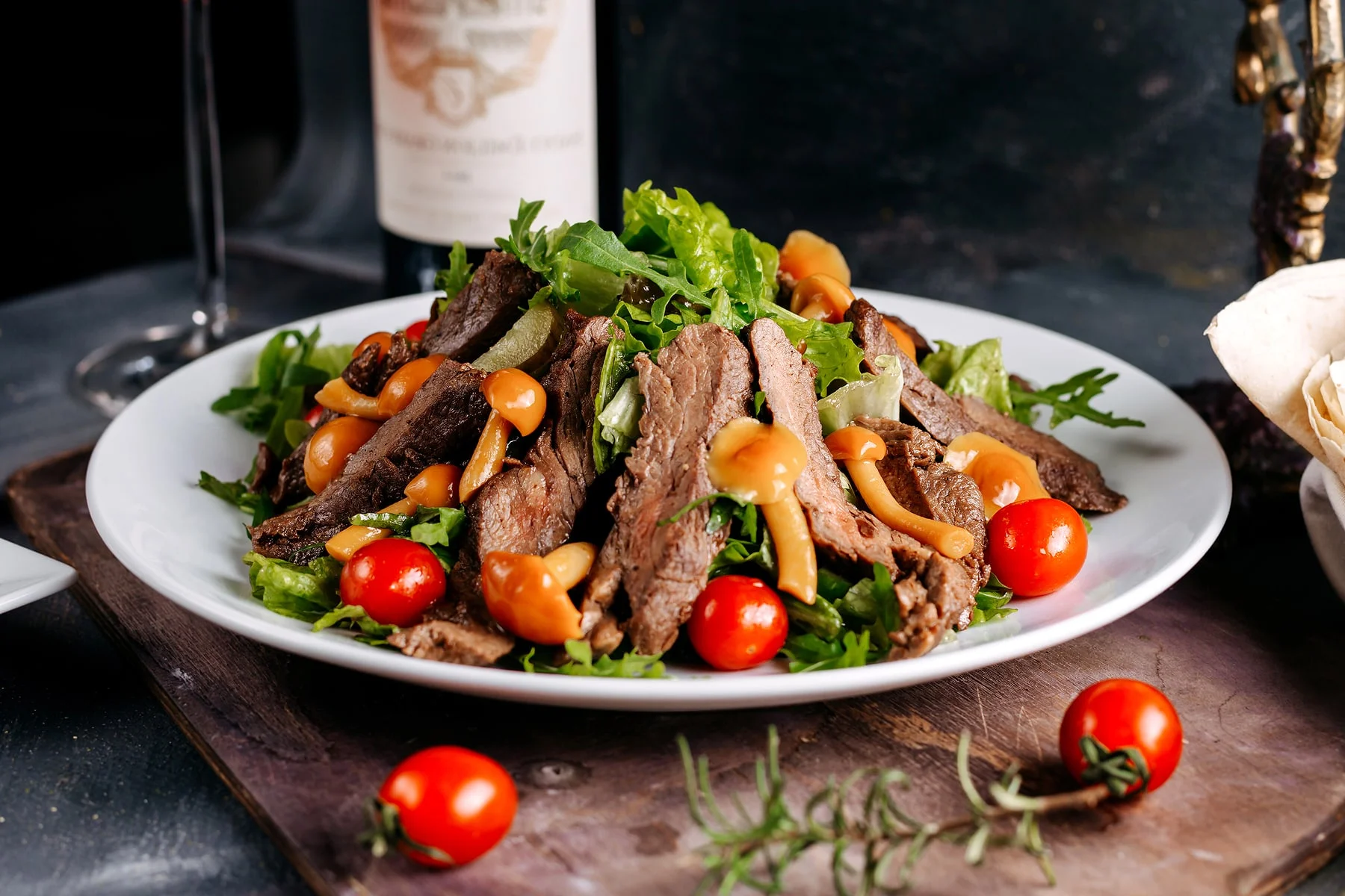Warm beef salad topped with marinated mushrooms, fresh greens, and cherry tomatoes on a white plate.