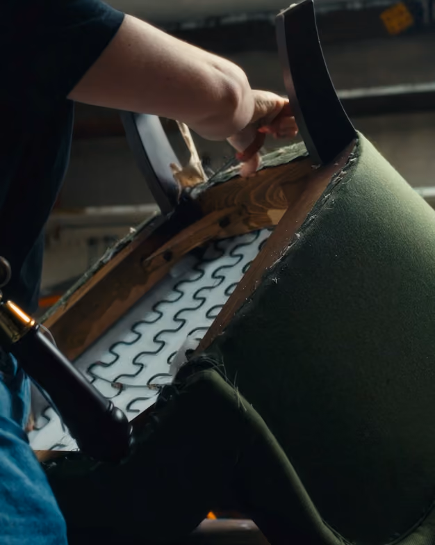 Person upholstering a green fabric chair, showing the chair's springs and wooden frame.