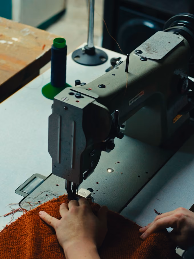 Hands guiding orange fabric through an industrial sewing machine on a white table with thread spool.
