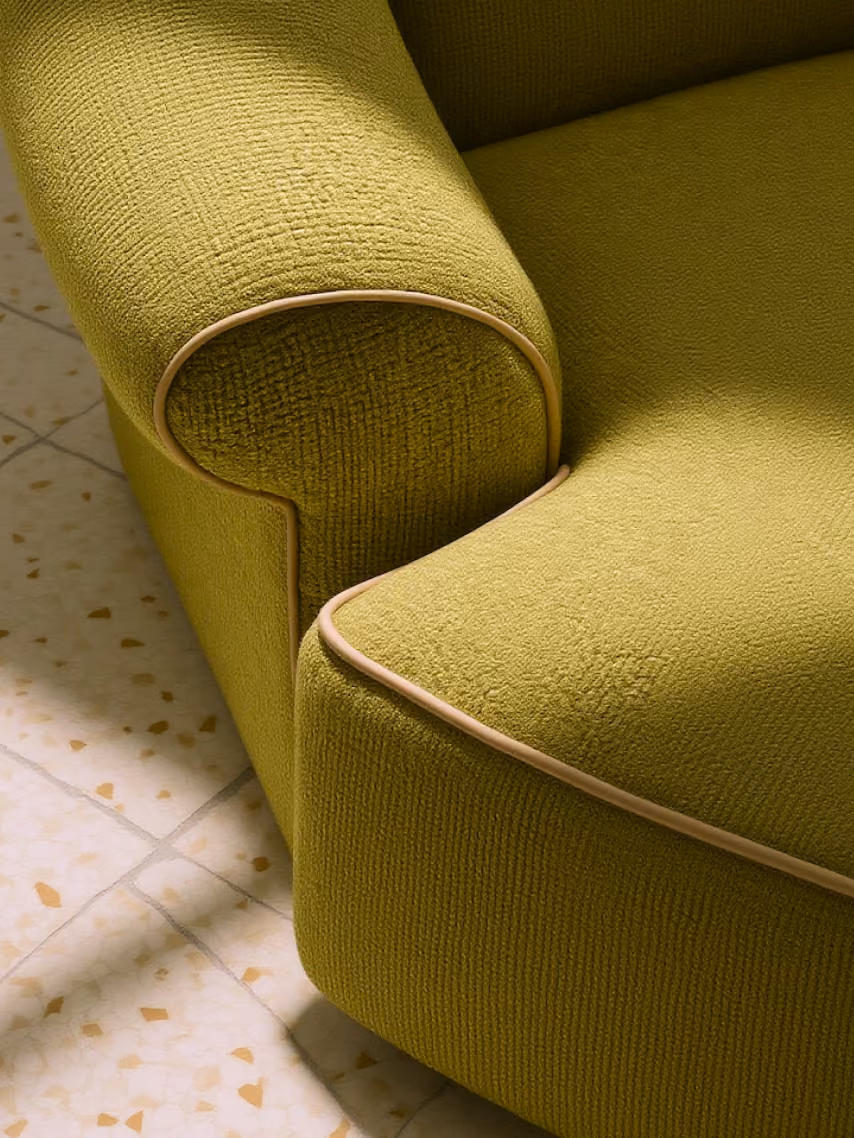 Close-up of a green textured upholstered sofa arm and seat with beige piping on a tiled floor.
