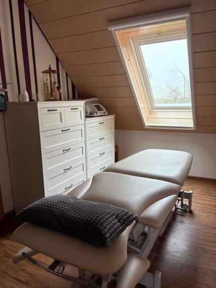 Behandlungszimmer mit verstellbarer Liege, Kissen, weißen Schränken und Dachfenster.