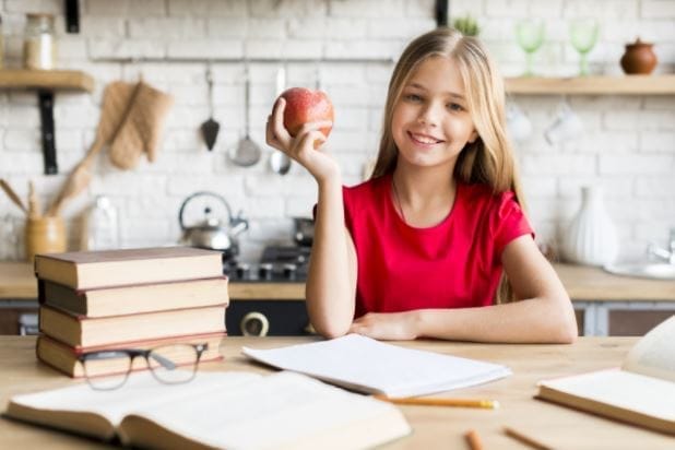 Mädchen sitzt lächelnd mit einem Apfel an ihrem Schreibtisch mit Büchern