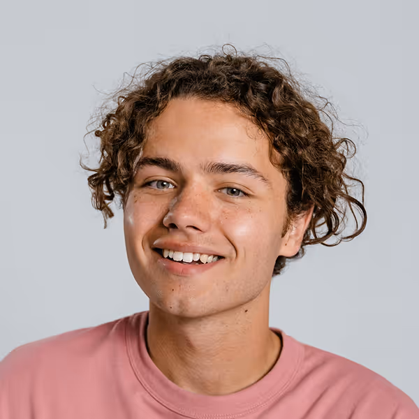 Young man with curly brown hair smiling and wearing a pink crewneck shirt against a plain gray background.