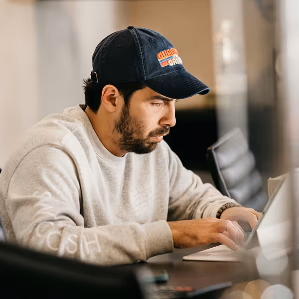 Man in a dark cap and gray sweatshirt focused on typing on a laptop at a table indoors.