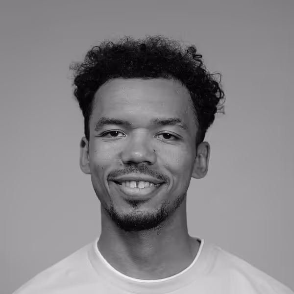 Smiling man with curly hair and facial hair wearing a light-colored shirt against a plain background.