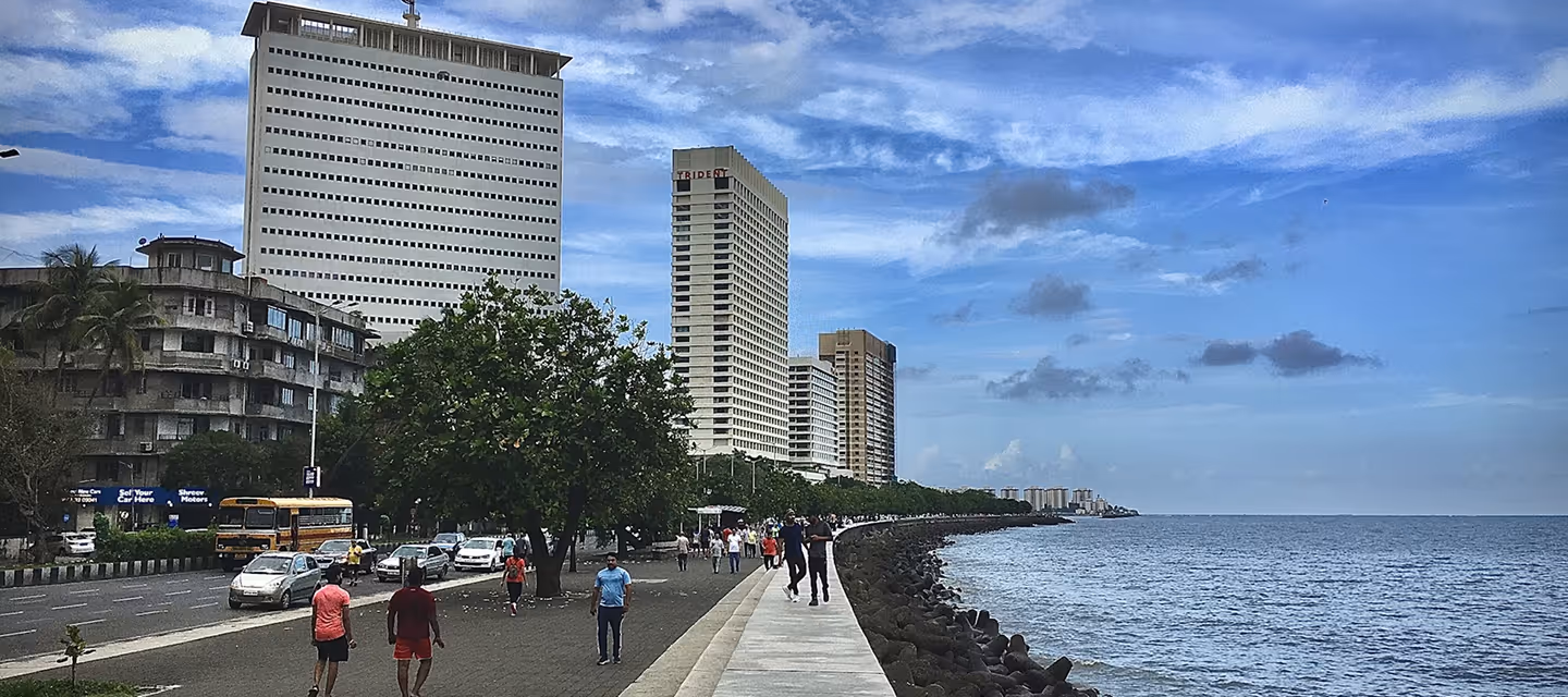 People walking along a waterfront promenade with tall buildings and a blue sky.