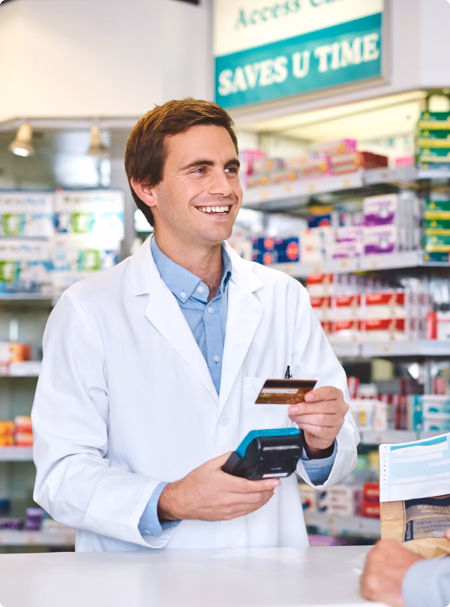 Smiling pharmacist in white coat holding a payment terminal and credit card in a pharmacy.