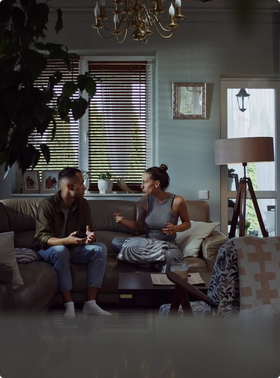 A man and woman sitting on a couch having a serious conversation in a living room.