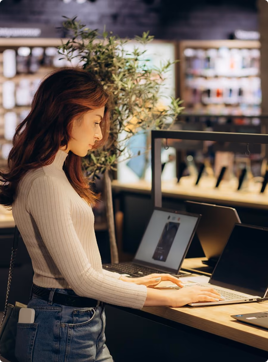 Woman in a cream sweater uses a laptop at a wooden counter in a tech store.