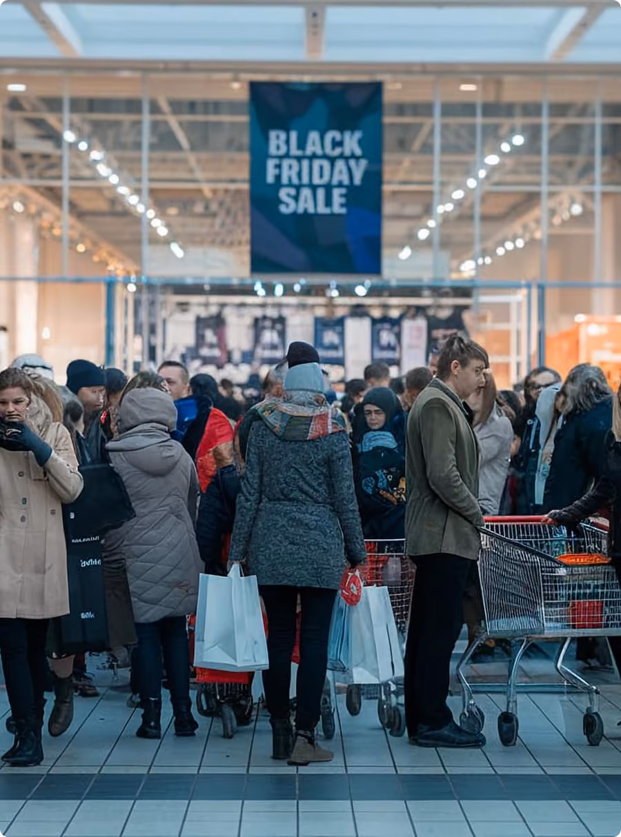 Crowded store entrance with people carrying bags and carts under a large Black Friday Sale banner.