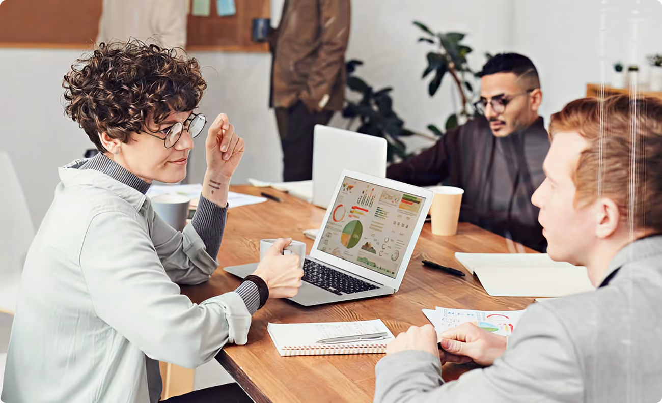Three people discussing data charts on laptops and papers at a wooden meeting table in an office.