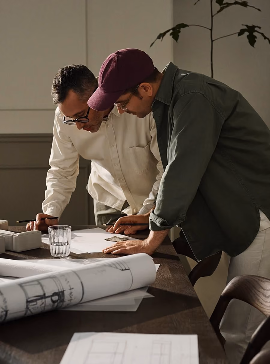 Two men leaning over a table reviewing architectural blueprints and samples in a workspace.