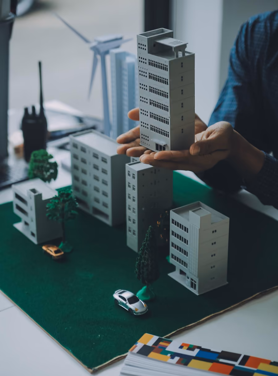 Hands holding a miniature gray building over a model city with buildings, trees, and cars.