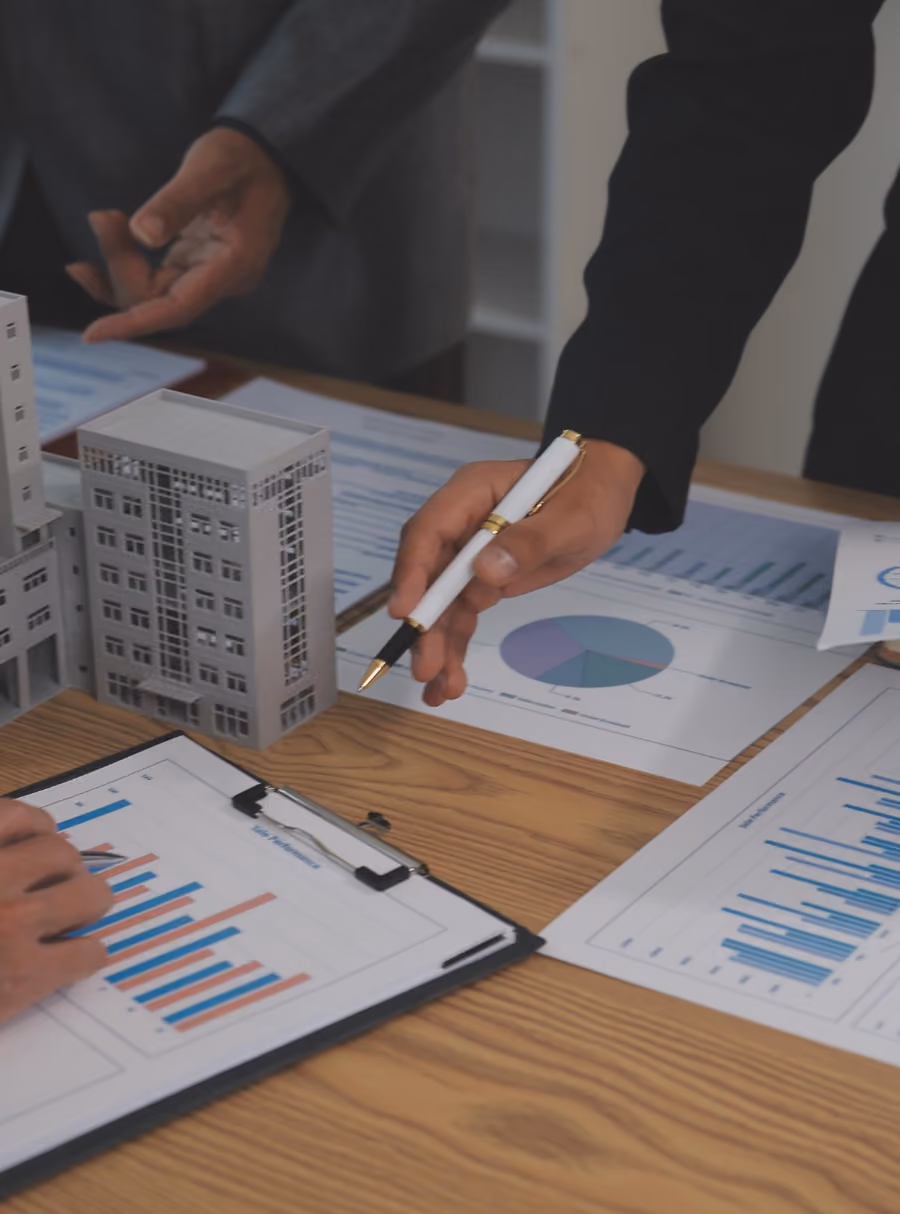 Hands with a pen pointing at charts near architectural building models on a wooden table.