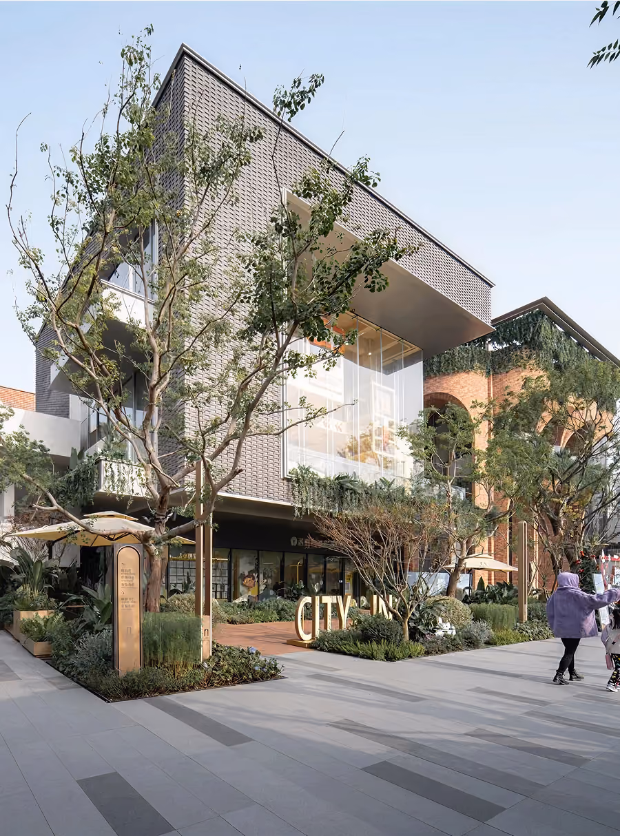 Modern two-story building with large windows and greenery in an outdoor urban plaza with people walking.