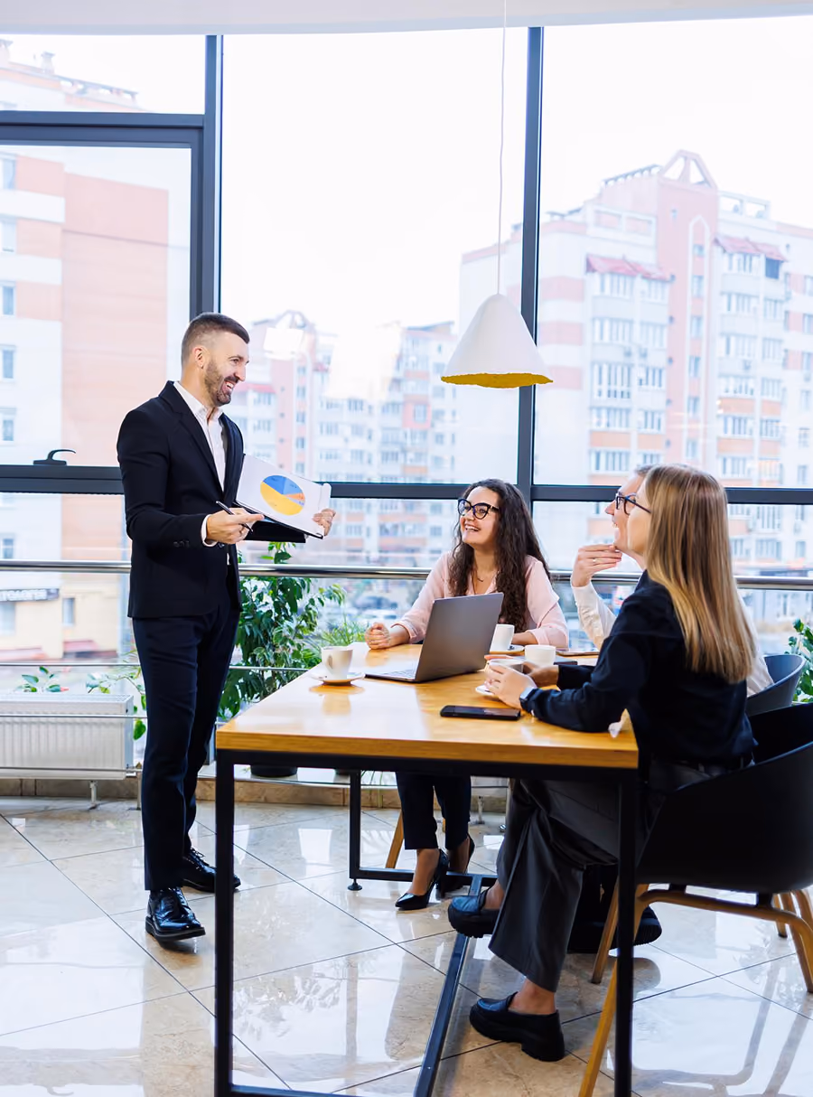 Businessman presenting pie chart to three colleagues seated at a table with a laptop in a bright office.