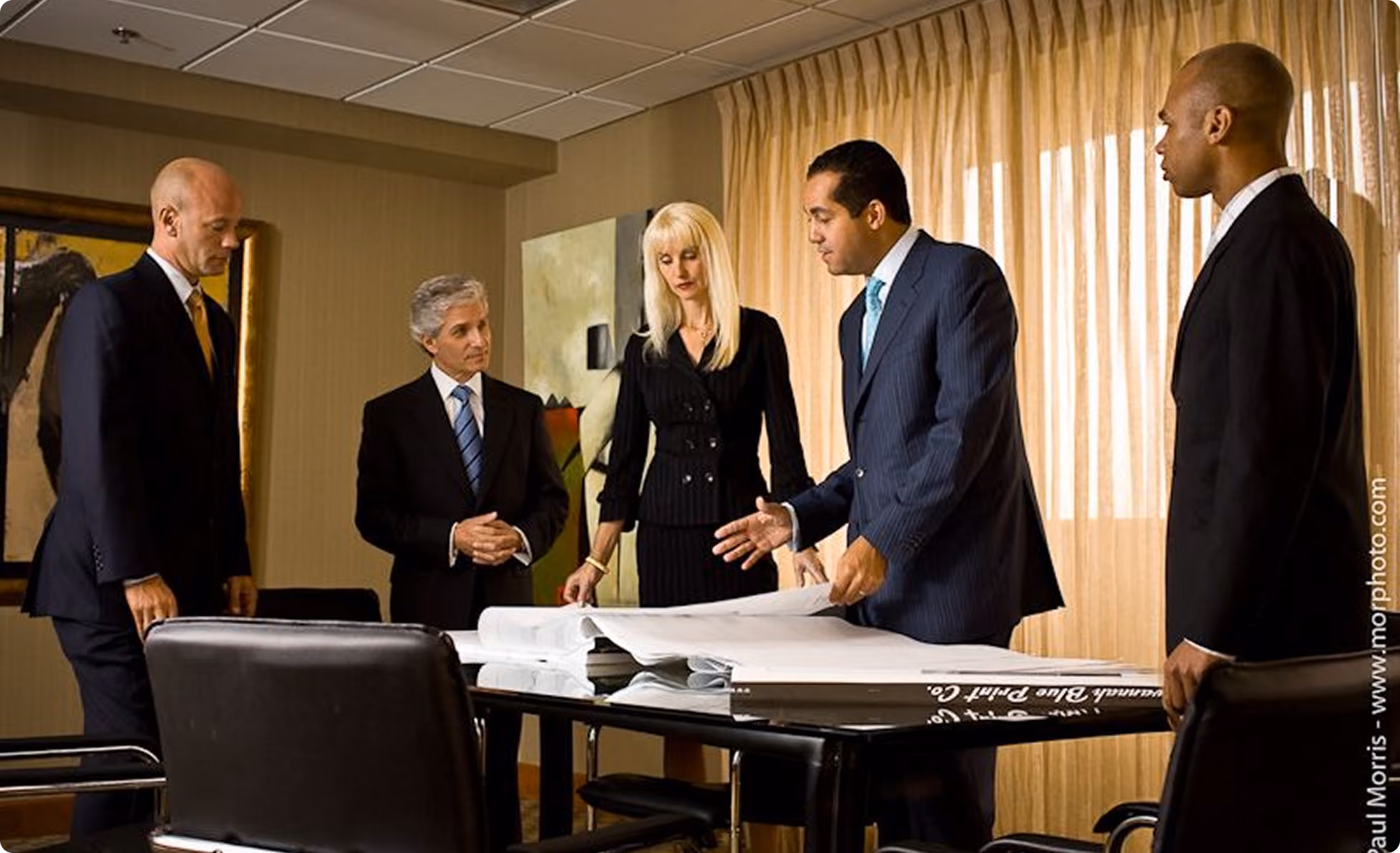 Five business professionals in suits gathered around a table reviewing large architectural plans.