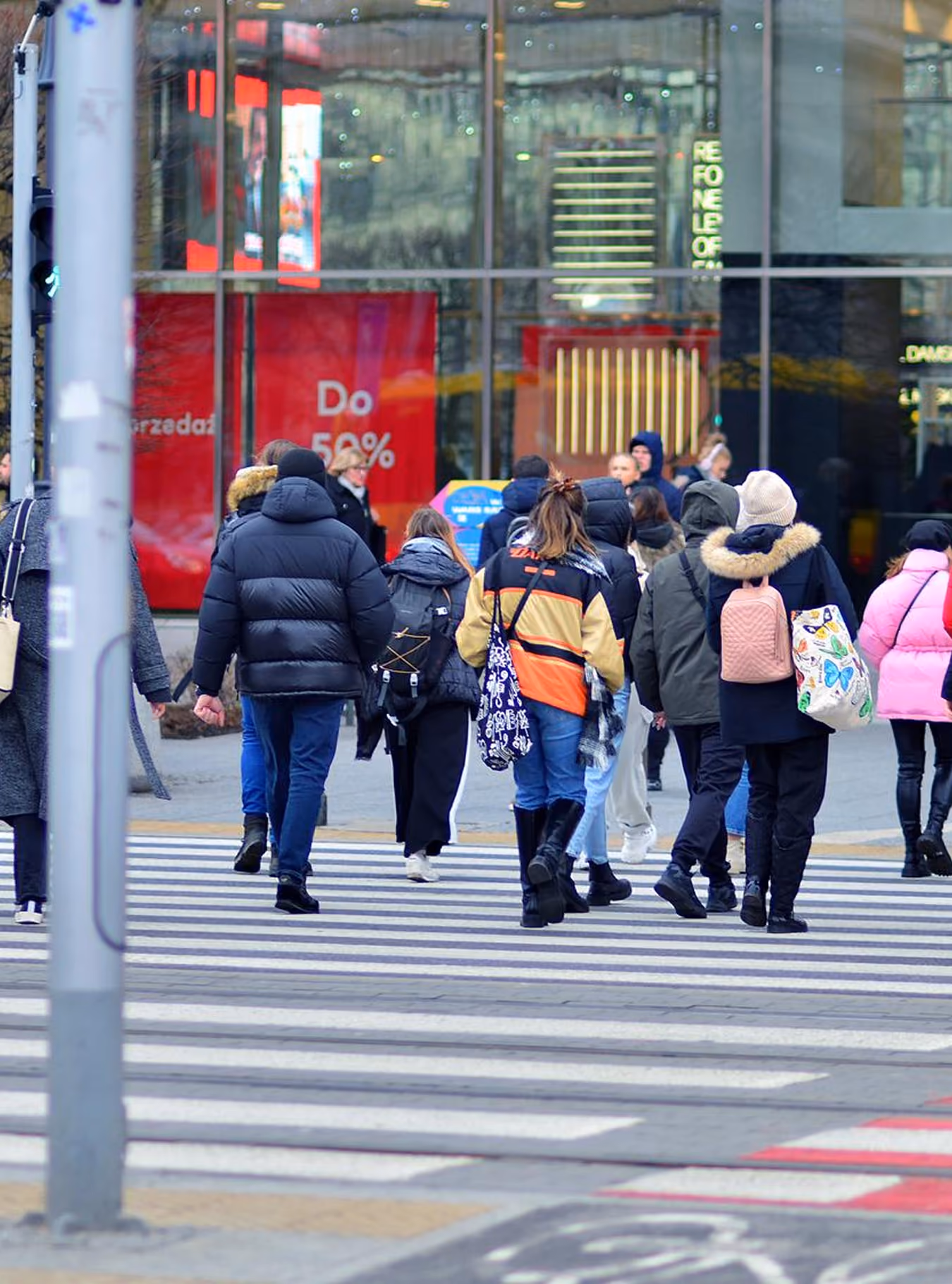 People wearing winter clothes walking across a city crosswalk in front of a glass building with red signs.