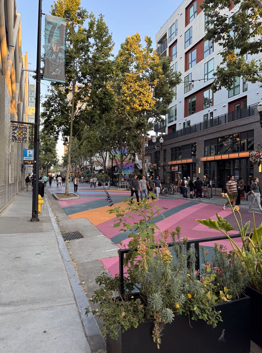 Urban street scene with colorful patterned pavement, pedestrians, trees, and modern buildings at sunset.