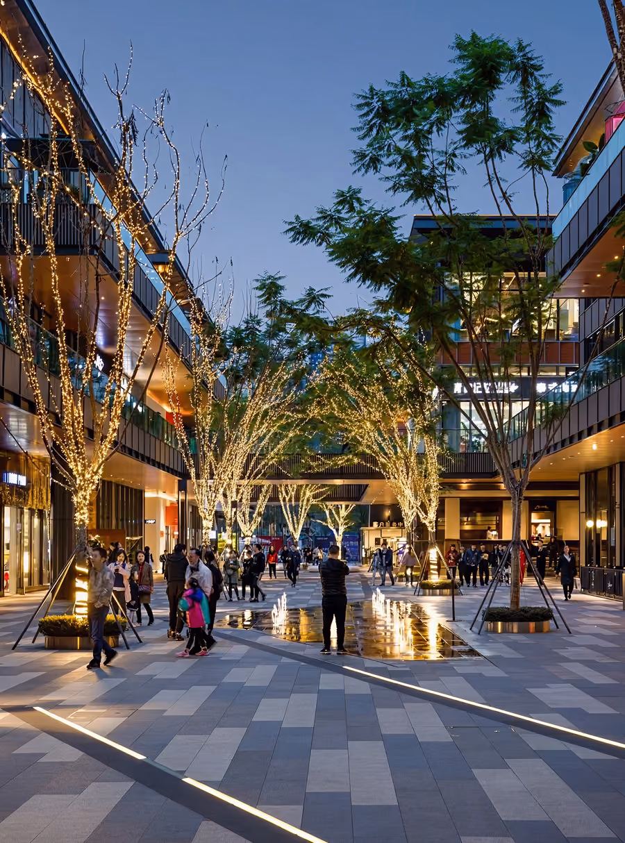 Evening city plaza with trees wrapped in lights and people walking along a tiled path and water fountains.