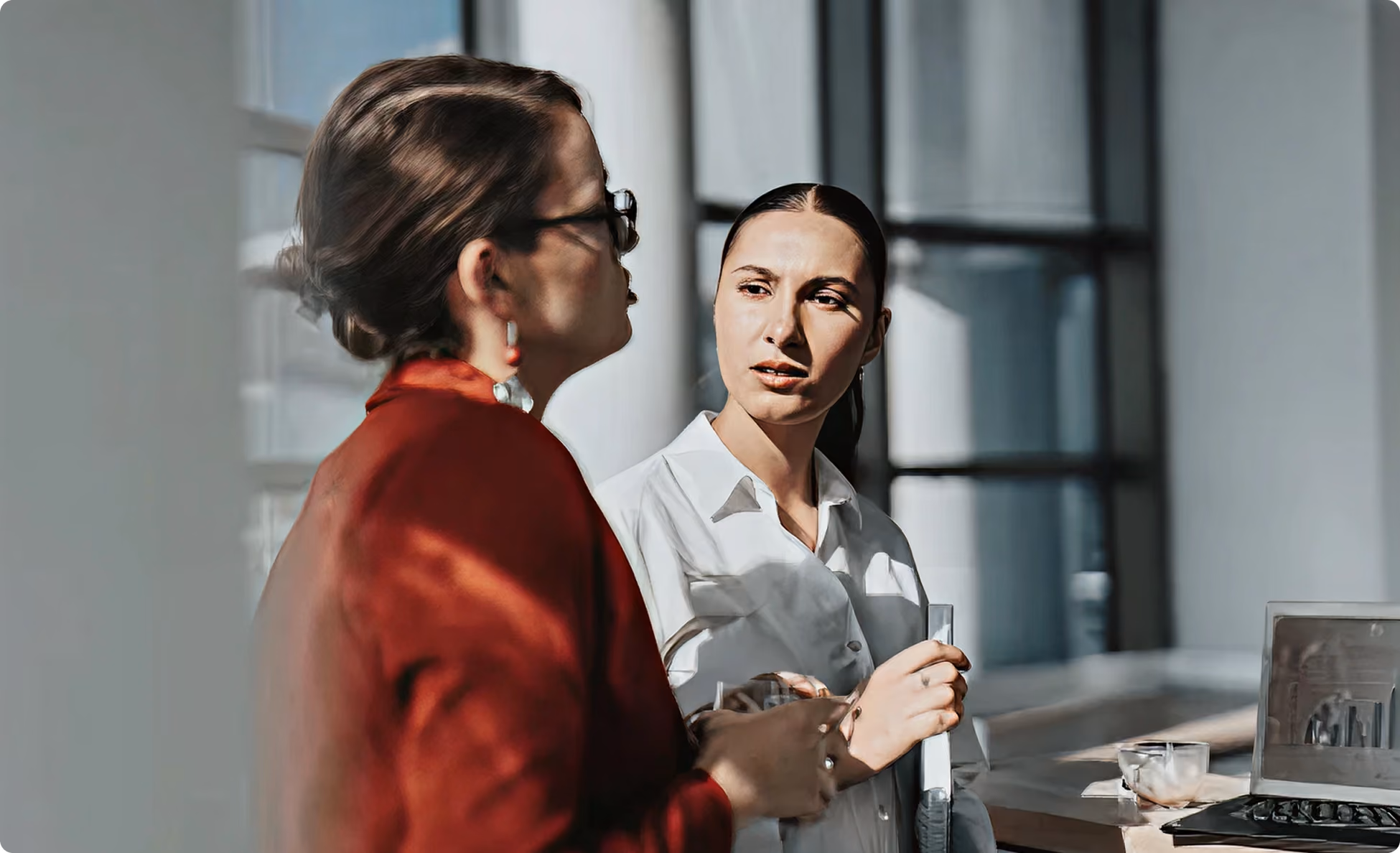 Two professional women engaged in conversation in a modern office with a laptop nearby.