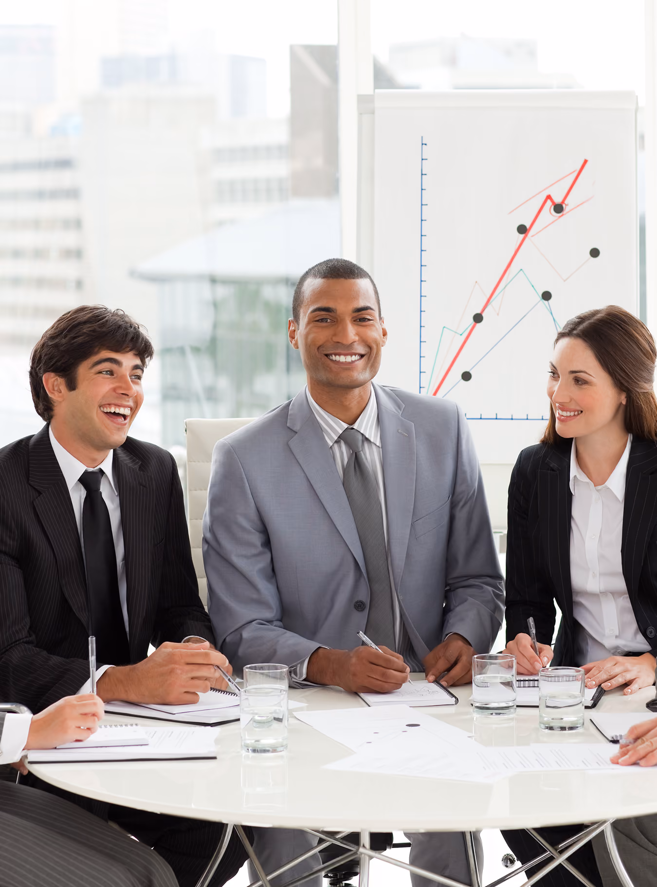 Three businesspeople in suits smiling and taking notes in a bright meeting room with graph behind them.