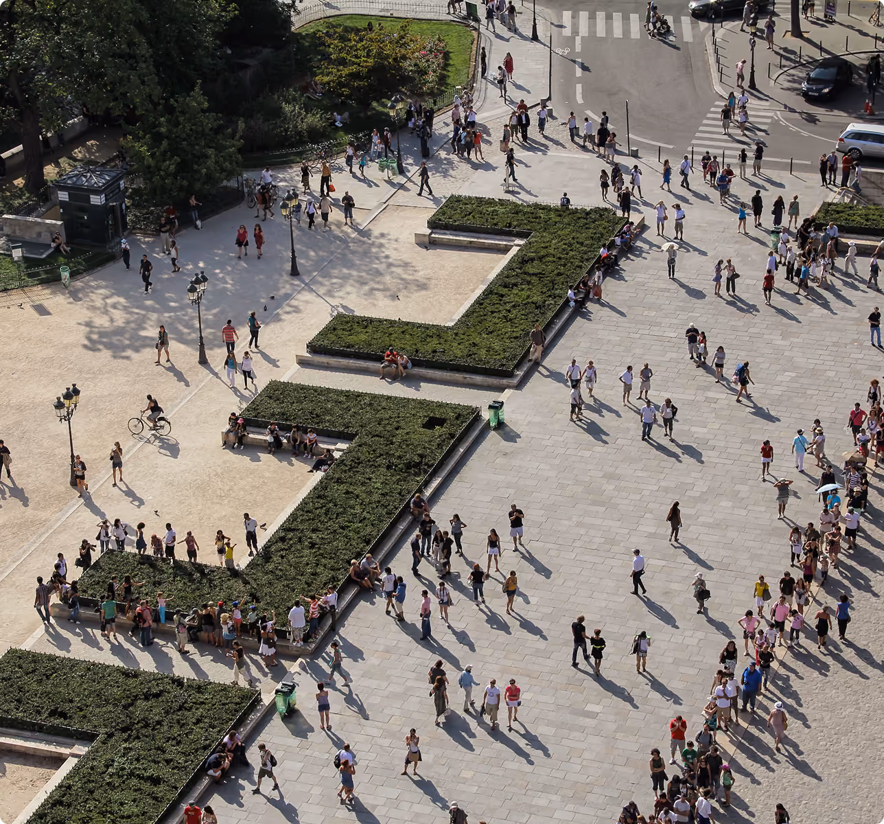 A large number of people walking and sitting near green hedges in a sunny open plaza with crosswalks.