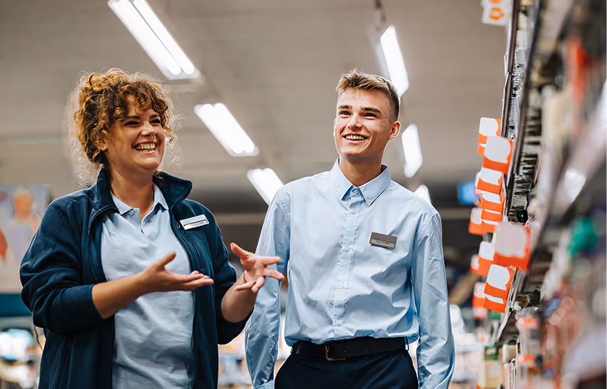Two young supermarket employees laughing and talking while standing by store shelves.