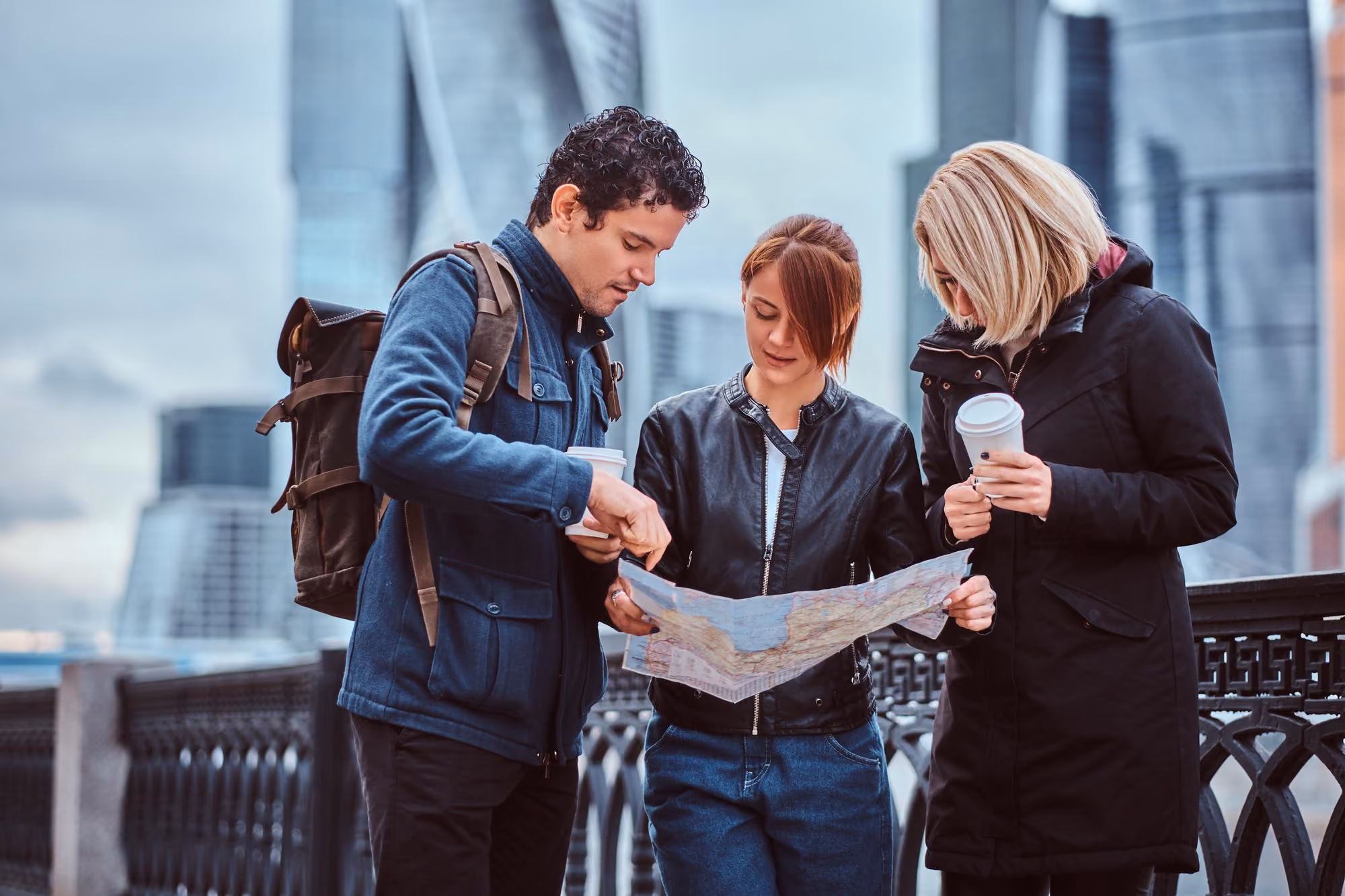 Three people in jackets looking at a map outdoors, with two holding coffee cups in a city setting.