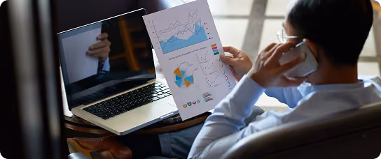 Person sitting with laptop, holding a sheet with charts, talking on a phone.