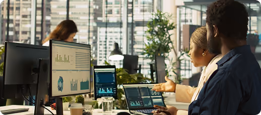 Two coworkers review data charts on laptop and monitors in an office with large windows.