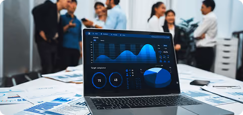 Laptop displaying data charts and graphs on desk with papers, with blurred people talking in background.