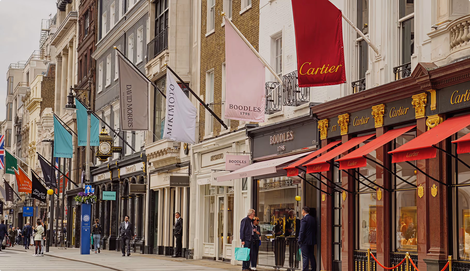 Street view of luxury jewelry stores with Cartier, Boodles, Mikimoto, Tiffany flags and pedestrians.