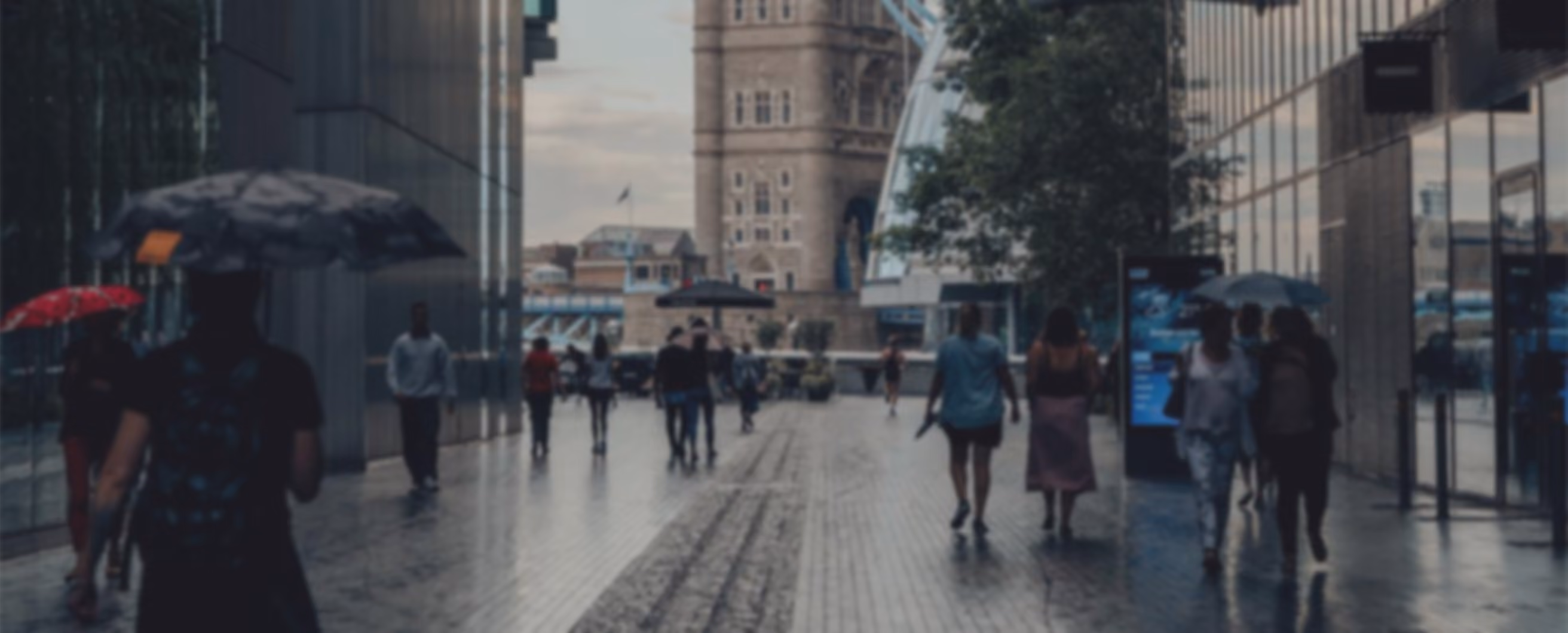 People walking on a wet city street under umbrellas near Tower Bridge on a cloudy day.