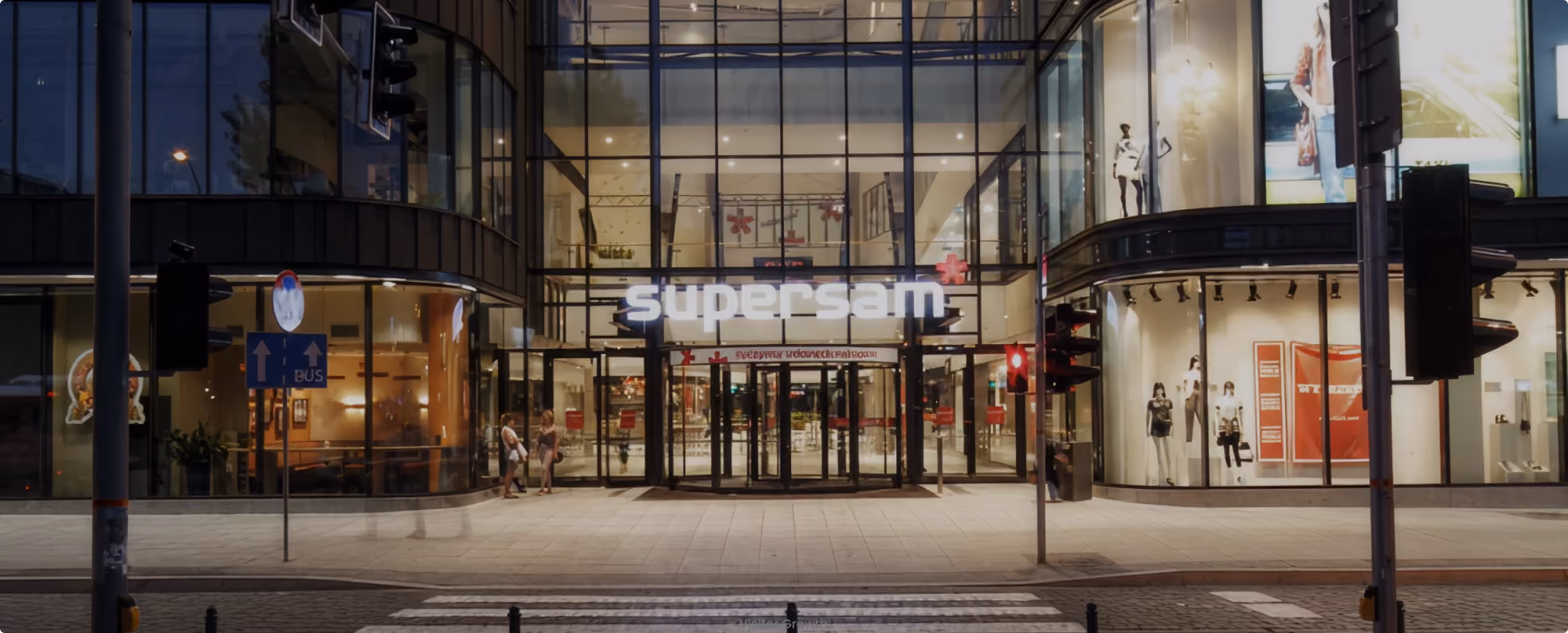 Entrance to a modern glass building with a lit sign reading 'supersam' and mannequins in display windows.