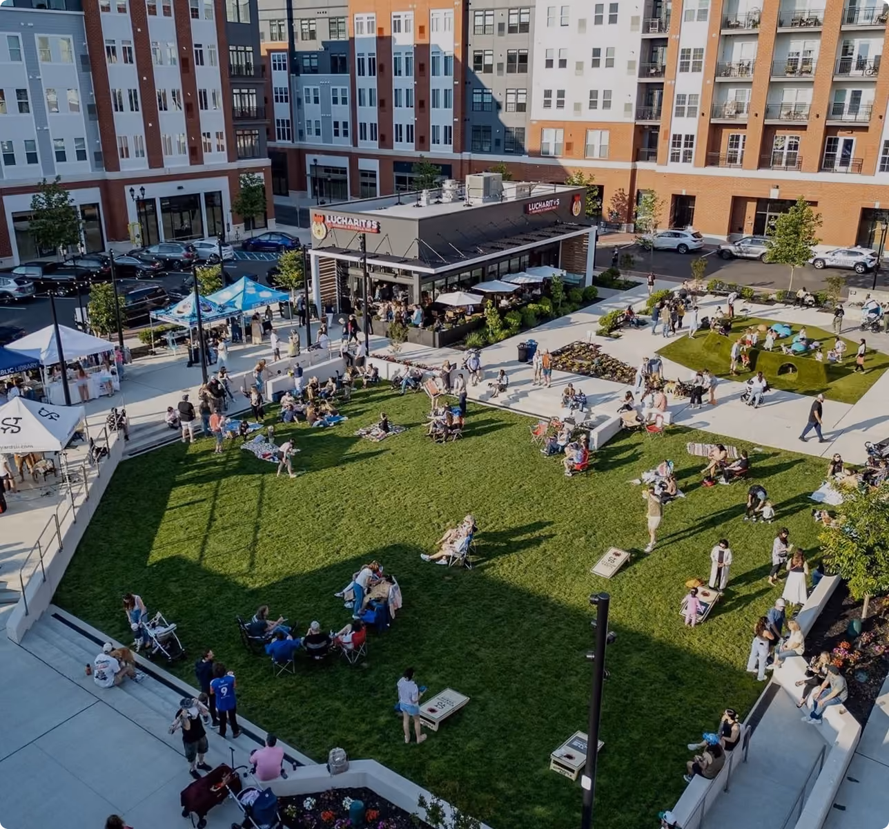 People relaxing on grass and walking near a restaurant in a courtyard surrounded by apartments.