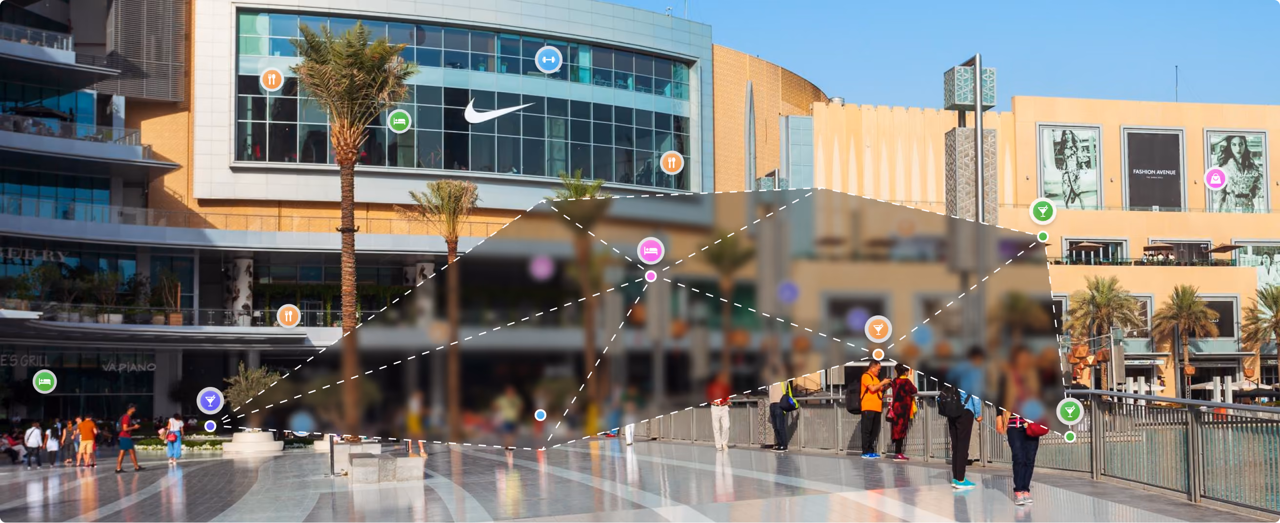 Outdoor shopping mall plaza with people walking and standing near buildings with stores and palm trees.